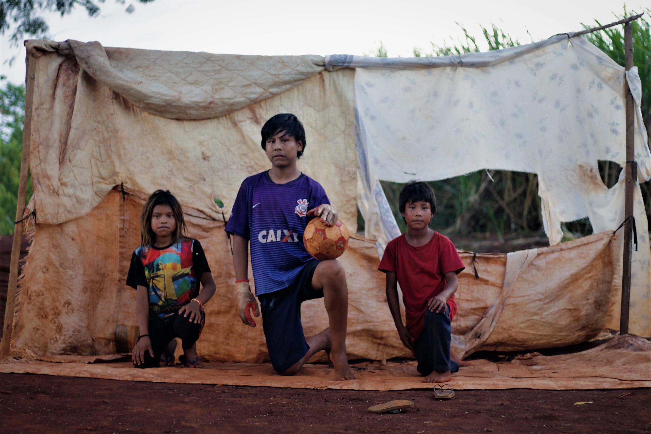Da esquerda para a direita, a neta e dois netos de Damiana posam para foto após brincarem de gol a gol, única diversão possível no pequeno terreno à margem da rodovia, em Mato Grosso do Sul. A criança do meio é o neto criado como filho por ela – o pai da criança morreu atropelado.