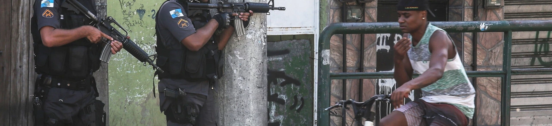 RIO DE JANEIRO, BRAZIL - NOVEMBER 20: Military Police (PM) officers hold their weapons as a young man bikes past in the Cidade de Deus 'City of God' favela community during an ongoing police operation on November 20, 2016 in Rio de Janeiro, Brazil. Seven local young men were found killed in the area today following a prolonged firefight between police and suspected gang members. In addition, four police officers were killed yesterday when their support helicopter crashed nearby during the firefight. According to preliminary indications the helicopter was likely not shot down. (Photo by Mario Tama/Getty Images) RIO DE JANEIRO, BRAZIL - NOVEMBER 20: Military Police (PM) officers hold their weapons as a young man bikes past in the Cidade de Deus 'City of God' favela community during an ongoing police operation on November 20, 2016 in Rio de Janeiro, Brazil. Seven local young men were found killed in the area today following a prolonged firefight between police and suspected gang members. In addition, four police officers were killed yesterday when their support helicopter crashed nearby during the firefight. According to preliminary indications the helicopter was likely not shot down. (Photo by Mario Tama/Getty Images)